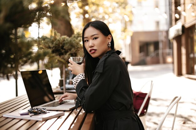 Attractive young brunette Asian woman in black stylish trench coat looks into camera drinks tea and works in laptop outside