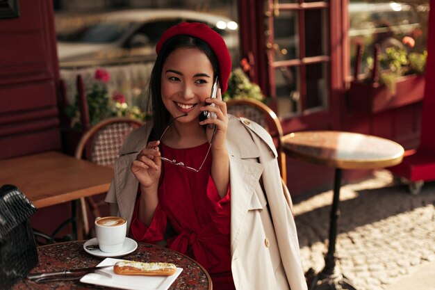 Attractive young brunette Asian woman in stylish trench coat red dress and beret smiles sincerely holds eyeglasses and talks on phone in street cafe