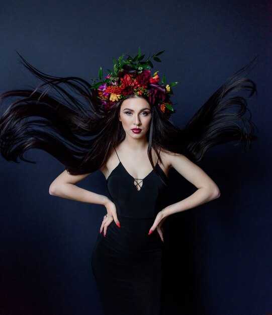 Attractive young brunette longhaired woman with bright make-up dressed in big colorful floral wreath on the dark blue background dressed in black dress