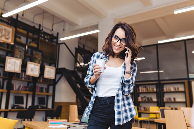 ライブラリに電話で話しているコーヒーと魅力的な若いブルネットの女性。コーヒーブレイク、大学生活、現代の仕事、勉強、賢い学生、いい仕事。