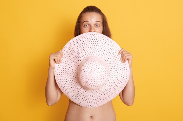 Attractive young brunette woman without clothes hide her boobs behind straw hat, sowing her bare belly, isolated over yellow wall.