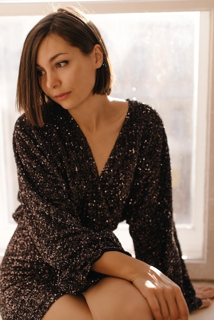 Привлекательная молодая кавказская брюнетка с короткой стрижкой в стильном черном платье сидит на подоконнике Концепция женской красоты