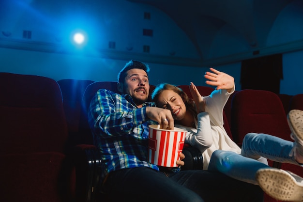 Attractive young caucasian couple watching a film at a movie theater, house or cinema. Look expressive, astonished and emotional. Sitting alone and having fun. Relation, love, family, weekend time.