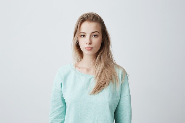 attractive young Caucasian dark-eyed female with long dyed blonde hair posing against gray blank wall dressed in casual blue sweater with calm face expression.