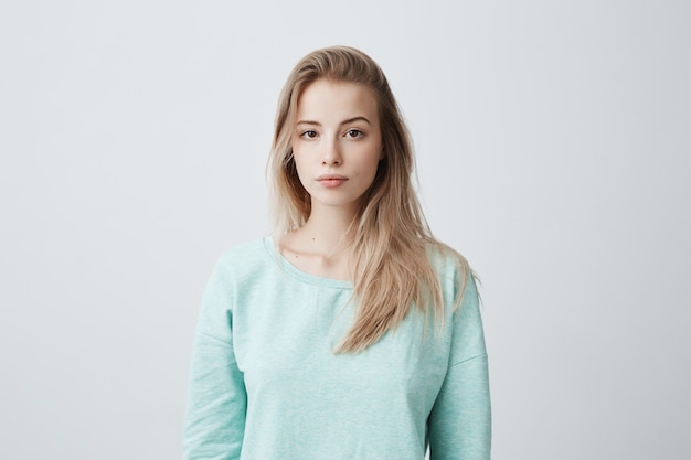 attractive young Caucasian dark-eyed female with long dyed blonde hair posing against gray blank wall dressed in casual blue sweater with calm face expression.