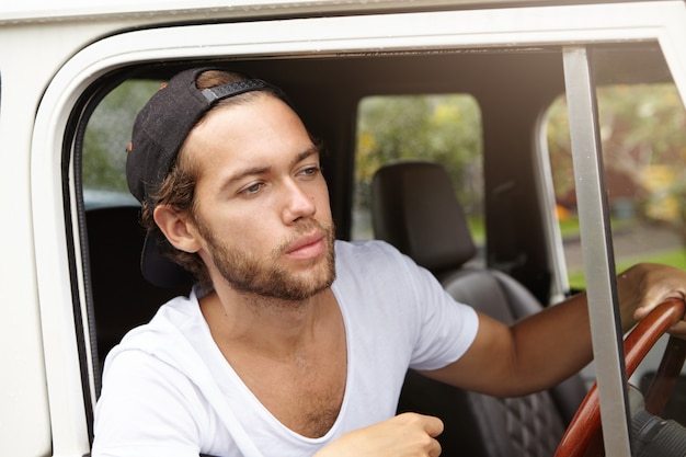 Attractive young Caucasian hipster man with beard wearing black baseball cap and white V-neck shirt driving white sport utility vehicle along country road