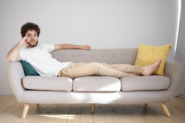 Attractive young Caucasian man with wavy hairdo and thick beard resting at home after work, lying barefooted on couch in his bachelor apartment, having pensive thoughtful expression,