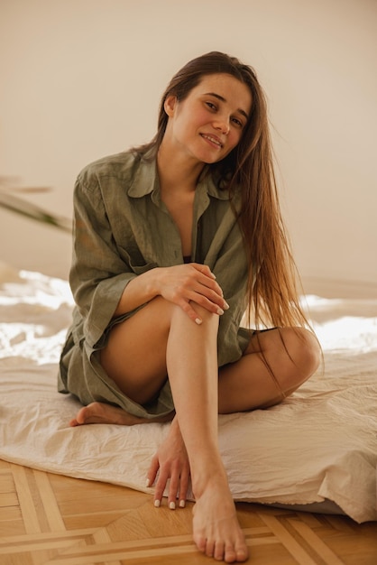 Attractive young caucasian woman sitting barefoot on mattress looking at camera with carefree smile Brunette woman with long hair wears casual clothes Leisure comfort and people