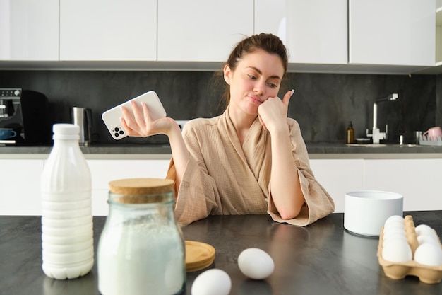 Free photo attractive young cheerful girl baking at the kitchen making dough holding recipe book having ideas