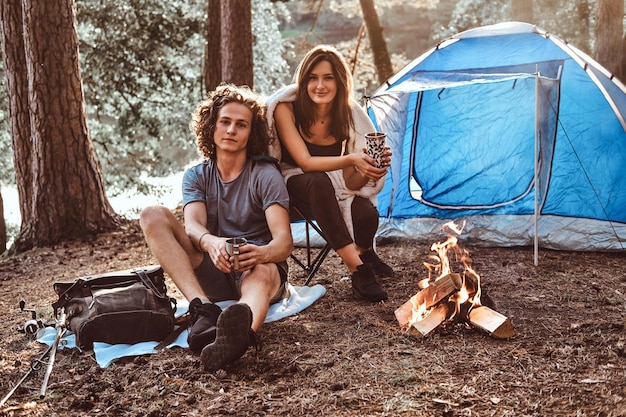 魅力的な若いカップルは、夏の森で一休みします。人々はテントとたき火の近くに座っています。