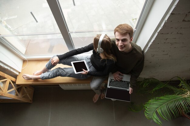 Attractive young couple using devices together, tablet, laptop, smartphone, headphones wireless. Communication, gadgets concept. Technologies connecting people in self-insulation. Lifestyle at home.