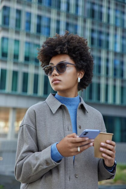 Attractive young curly haired woman wears sunglasses and stylish jacket uses cellphone chats in social networks drinks coffee poses outdoors near modern building People lifestyle and technology