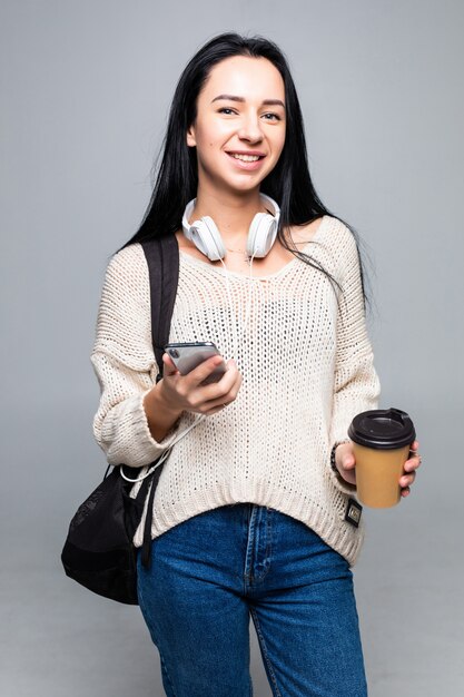 Attractive young cute brunette woman while using smart phone and drinking coffee isolated on gray wall