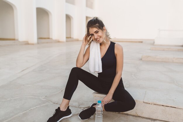 Attractive young cute woman relaxing after workout on white east architecture. Fitness, fashionable model, healthy lifestyle, motivation, true positive emotions, smiling