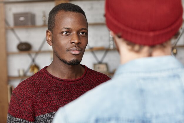 Attractive young dark-skinned man wearing casual sweater having talk to his unrecognizable friend that he came across at cafe, listening to him with happy interested look. Two men talking indoors