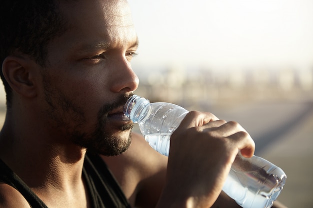 Attractive young dark skinned sportsman with short beard drinking water from bottle looking far away with thoughtful face expression, dressed in black sleeveless shirt, relaxing after morning run