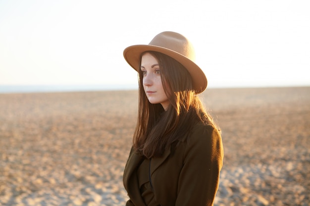 Attractive young European female dressed in stylish overclothes having nice walk along coastline on sunny day, came to the sea for contemplating sunset. Pretty woman in hat relaxing on sandy beach