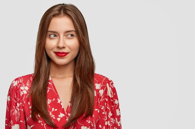 Attractive young European female with long dark hair, red painted lips and stylish blouse, looks pensively aside, has dreamy expression, stands against white wall with copy space on left side