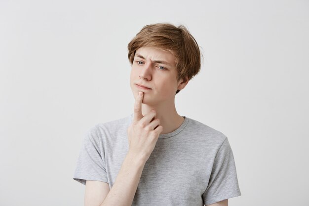 Attractive young fair-haired man with blue eyes dressed casually has thoughtful expression, keeps index finger on his chin, tries to recollect something or has great idea. Pensive male student