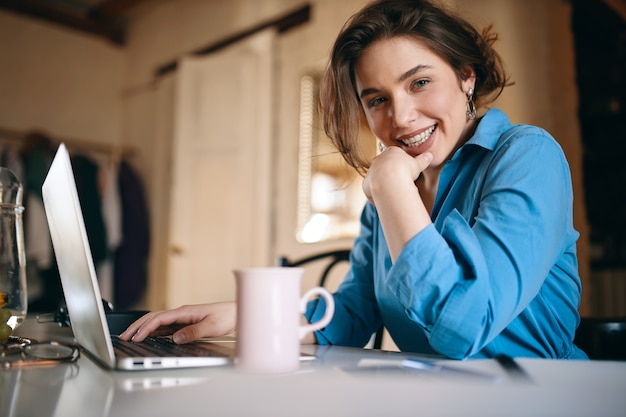 Foto gratuita attraente giovane designer femminile che lavora in remoto, aggiornando il sito web utilizzando il laptop.