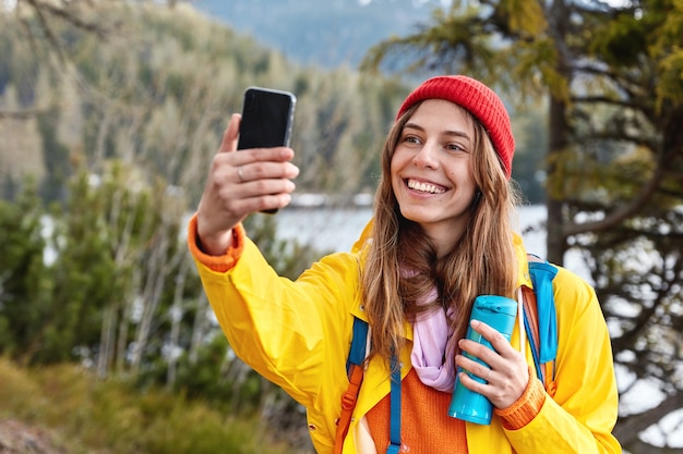 Attractive young female tourist makes selfie portrait on smart phone, drinks hot coffee or tea from thermos