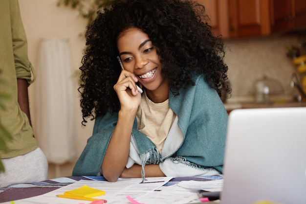 Attractive young female with Afro hairstyle abd braces having phone conversation and smiling happily while doing paperwork at home, sitting at kitchen table with lots of papers and laptop computer
