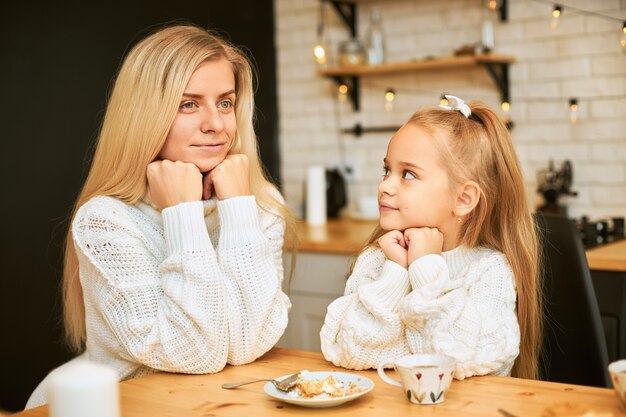 Attractive young female with blonde long hair and her beautiful daughter both in cozy sweaters having breakfast in kitchen sitting at dining table, drinking tea, having cake, keeping hands under chin
