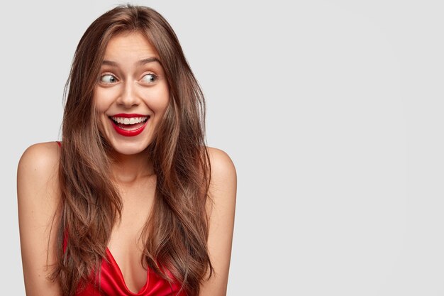 Attractive young female with long dark straight hair, has happy expression, red lips, dressed casually, stands against white wall