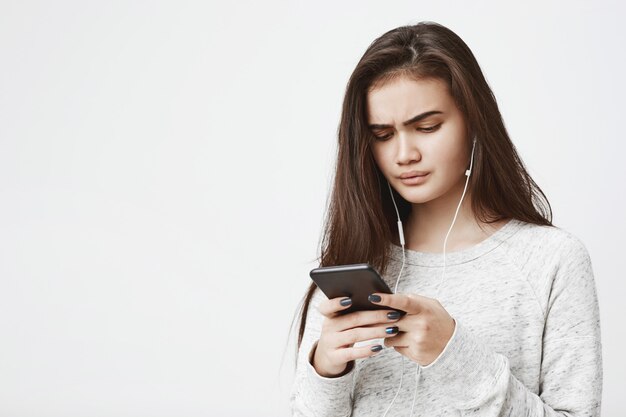 Attractive young female with worried expression holds phone and listen music