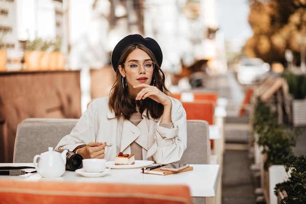 Привлекательная молодая девушка в классических бежевых очках плаща сидит за столом с чизкейком и чаем на террасе городского кафе солнечной осенью
