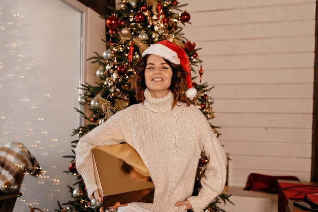 크리스마스 트리에 기대어 서 있는 동안 선물 상자를 들고 있는 매력적인 어린 소녀 Redhaired 여자가 스웨터와 산타 모자를 쓰고 있다