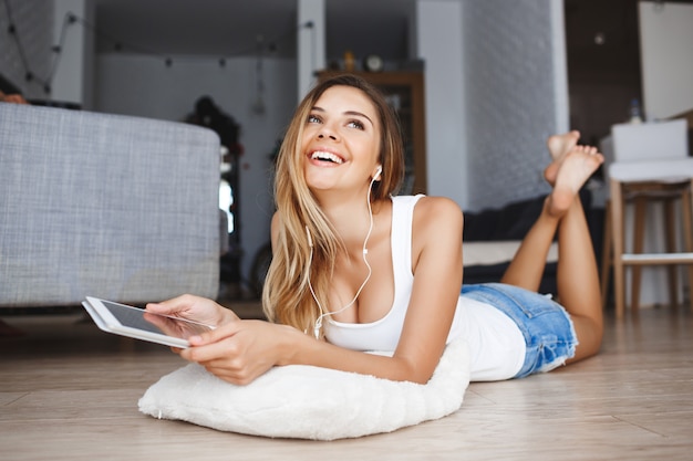 Attractive young girl laying on floor and holding tablet in hands with earphones at living room looking aside smiling