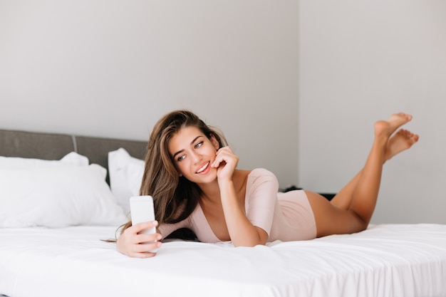 Attractive young girl taking selfie on phone on bed in apartment in the morning.