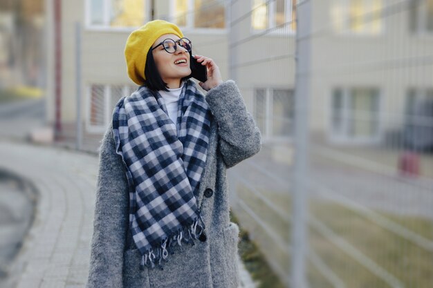 通りを歩いて、電話と笑顔で話しているコートでサングラスをかけている魅力的な若い女の子