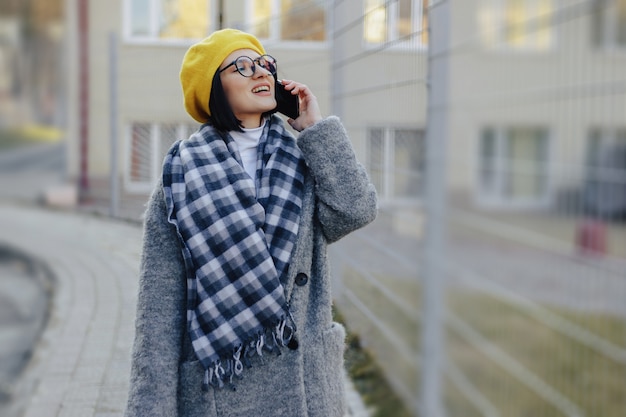 Привлекательная молодая девушка в темных очках в пальто гуляет по улице и разговаривает по телефону и улыбается