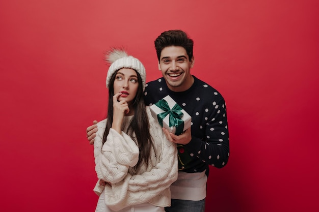 Attractive young girl with dark long hair bright lips light sweater waiting for surprise from gorgeous brunette in Christmas jersey holding gift box Posing isolated on red background