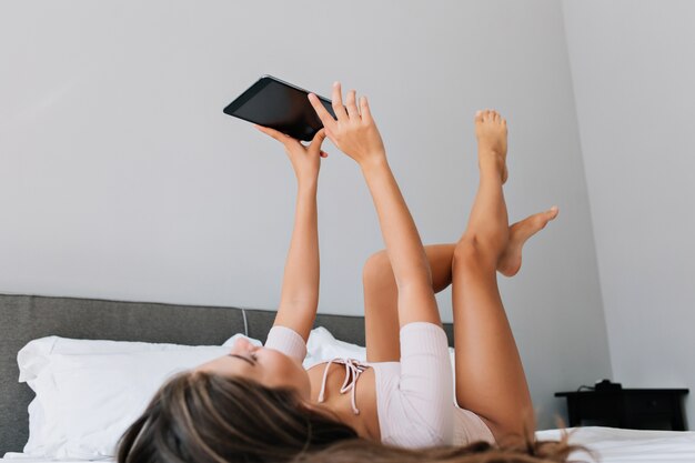 현대 아파트에서 침대에 재미 긴 머리를 가진 매력적인 젊은 여자. 그녀는 위에 태블릿과 장난 꾸러기 다리를 들고 있습니다.