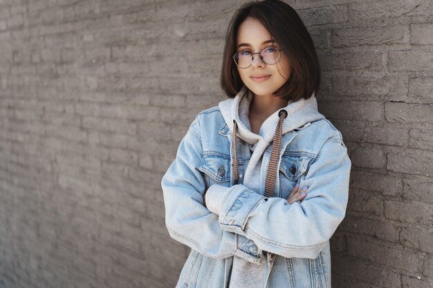 Attractive young girl with short hair, wearing glasses and street style clothes, leaning on a brick wall.