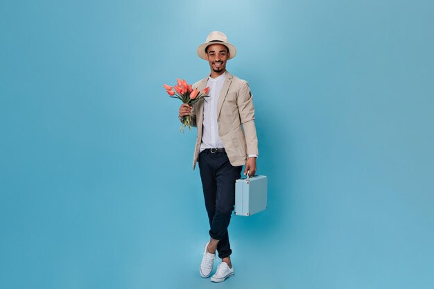 Привлекательный молодой парень в шляпе позирует с розовыми цветами и чемоданом