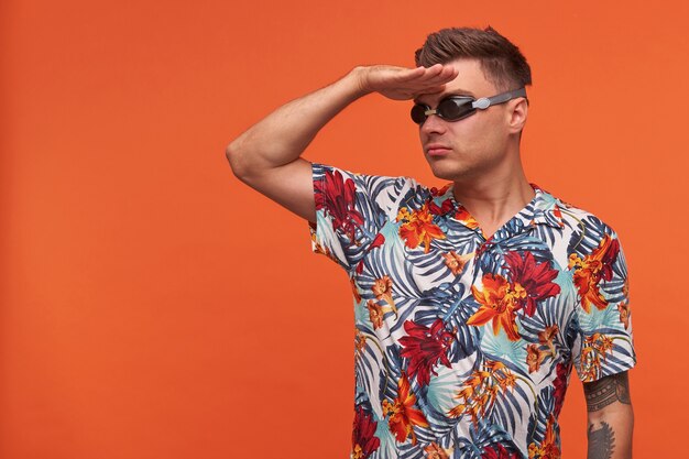Free photo attractive young guy posing like captain of ship, making patrol gesture, raising palm to forehead, wearing swimming glasses and flowered shirt
