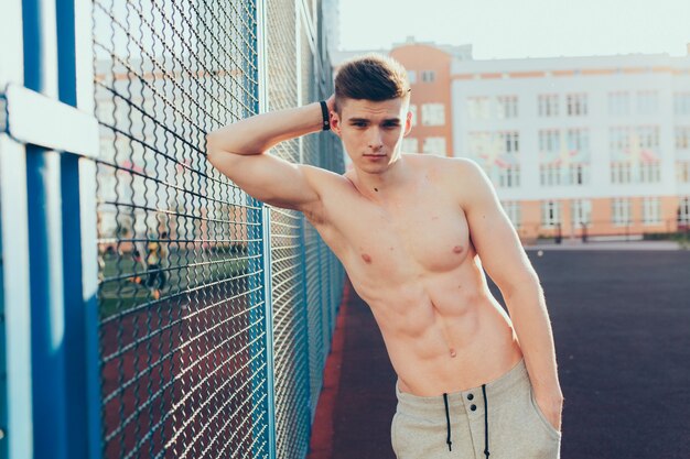 Attractive young guy with naked muscular torso posing near fence in the morning on stadium. He wears gray knickers. He looks to camera confidently.