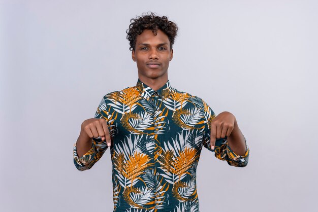 Привлекательный молодой красивый темнокожий мужчина с вьющимися волосами в рубашке с принтом листьев указывает вниз обоими указательными пальцами