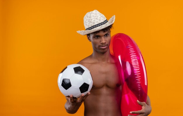 An attractive young handsome dark-skinned man with curly hair wearing sun hat holding inflatable pool ring and soccer ball 