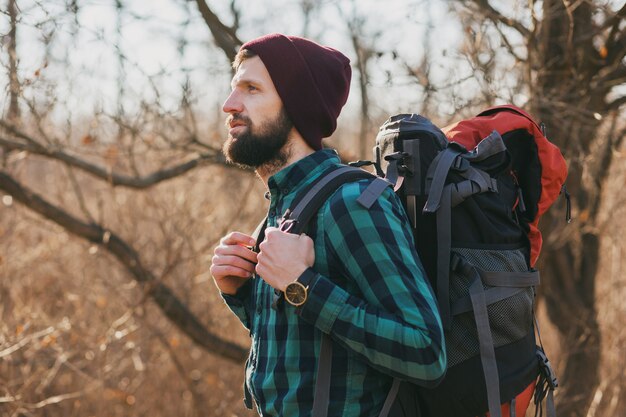 Attractive young hipster man traveling with backpack in autumn forest wearing checkered shirt and hat, active tourist, exploring nature in cold season