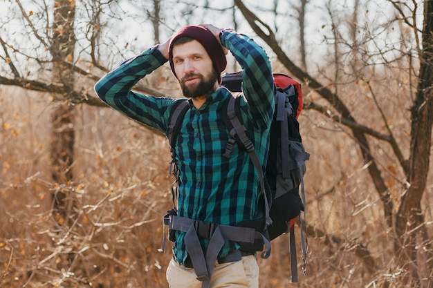 Привлекательный молодой хипстерский мужчина путешествует с рюкзаком в осеннем лесу в клетчатой рубашке и шляпе, активный турист, исследующий природу в холодное время года