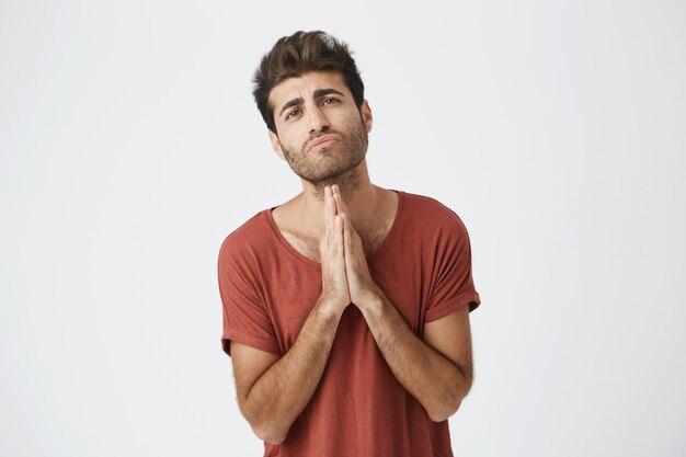 Attractive young italian male in red t-shirt holding palms pressed together in front of him with regretful expressions apologizing to girlfriend.