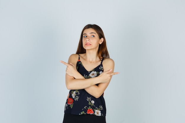 Attractive young lady in blouse pointing to the opposite sides and looking bewildered , front view.