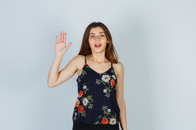 Attractive young lady in blouse waving hand for greeting and looking amazed , front view.