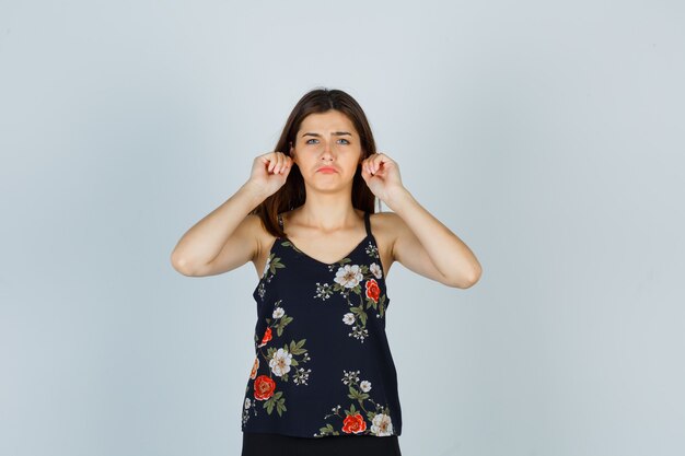 Attractive young lady pulling down her earlobes, curving lips, frowning face in blouse and looking sad , front view.