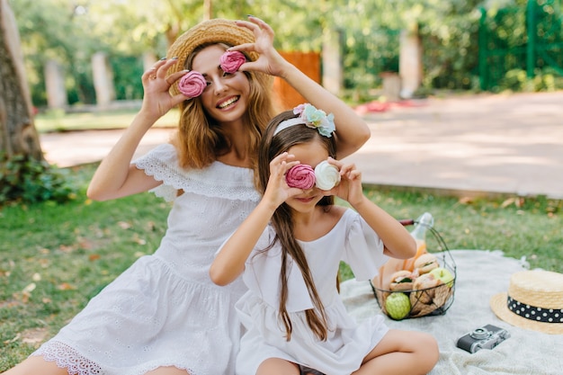 娘と冗談を言ったり、カラフルなクッキーで遊んでレトロな麦わら帽子の魅力的な若い女性。夏の公園でピクニックをして笑っている2人のかわいい姉妹。
